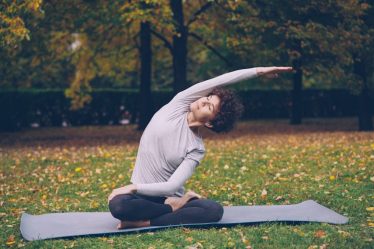 Slim young lady is doing sequence of yoga asanas sitting on mat in park alone on warm autumn day with beautiful grass and trees around her. Recreation and sport concept.