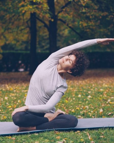 Slim young lady is doing sequence of yoga asanas sitting on mat in park alone on warm autumn day with beautiful grass and trees around her. Recreation and sport concept.