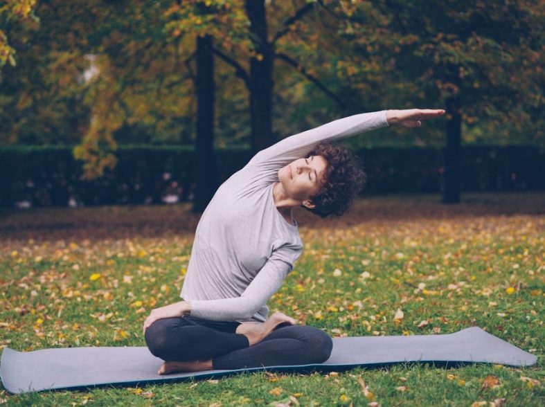 Slim young lady is doing sequence of yoga asanas sitting on mat in park alone on warm autumn day with beautiful grass and trees around her. Recreation and sport concept.